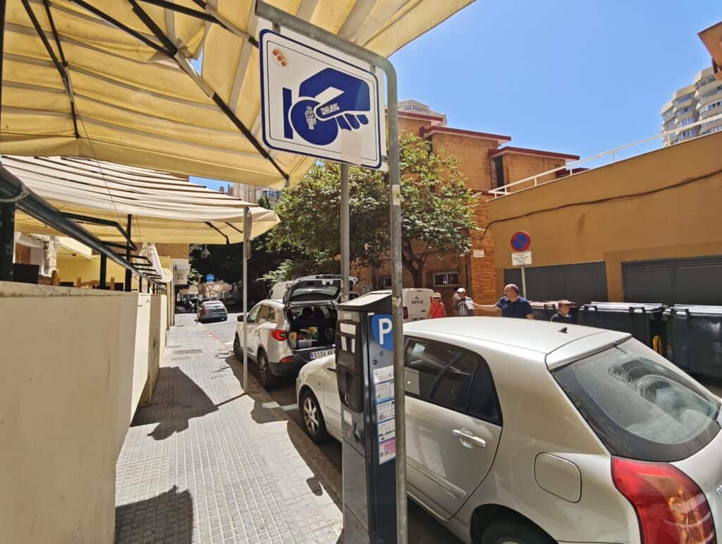 Handy parking meter near a sidewalk in Malaga, Spain, with cars and pedestrians on a sunny day.