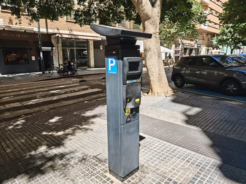 Parking meter onmalaga sidewalk with parked cars and trees in the background, typical urban scene.